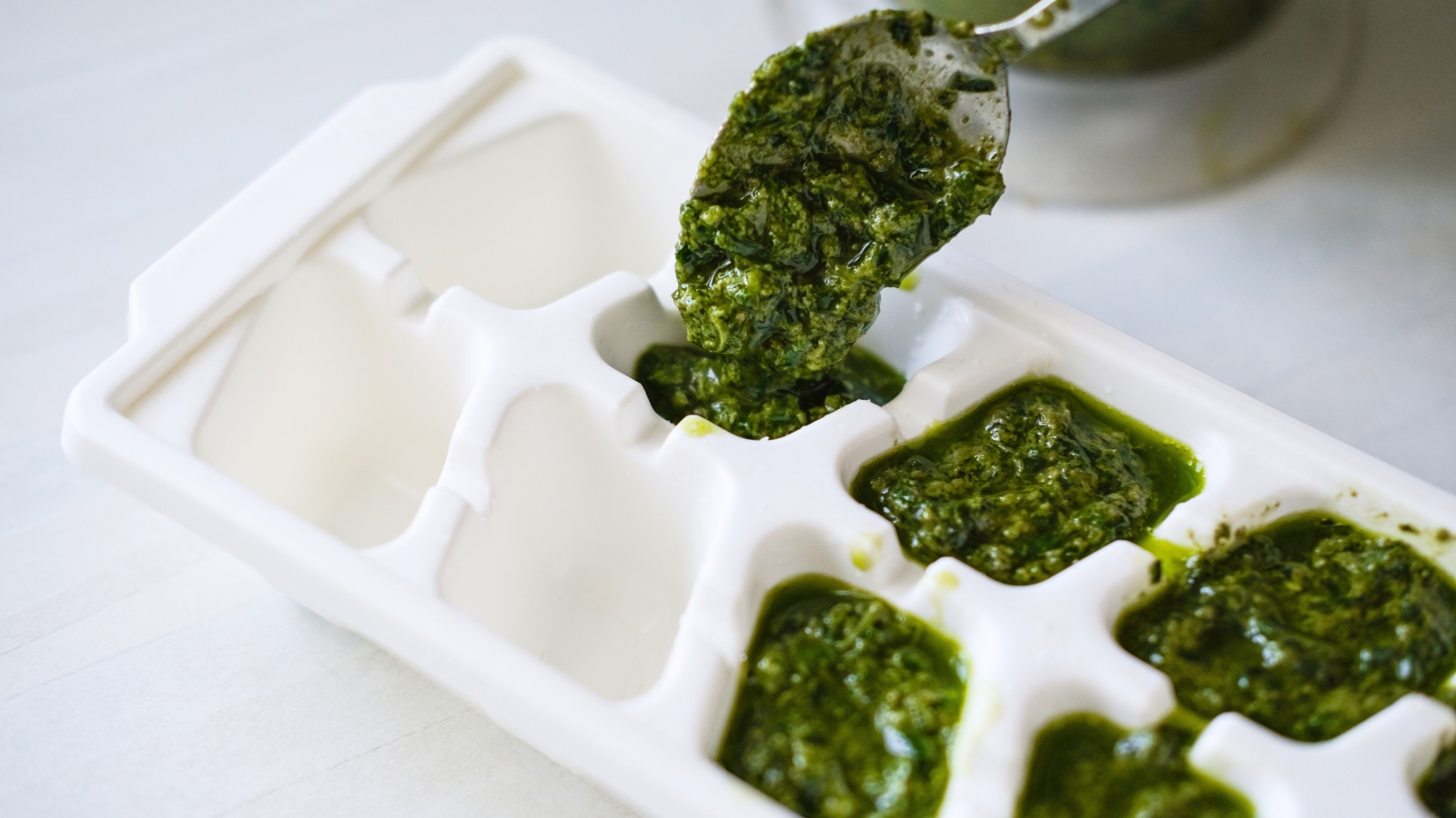 A white ice cube tray is being filled with vibrant green pesto using a spoon, ready for freezing.