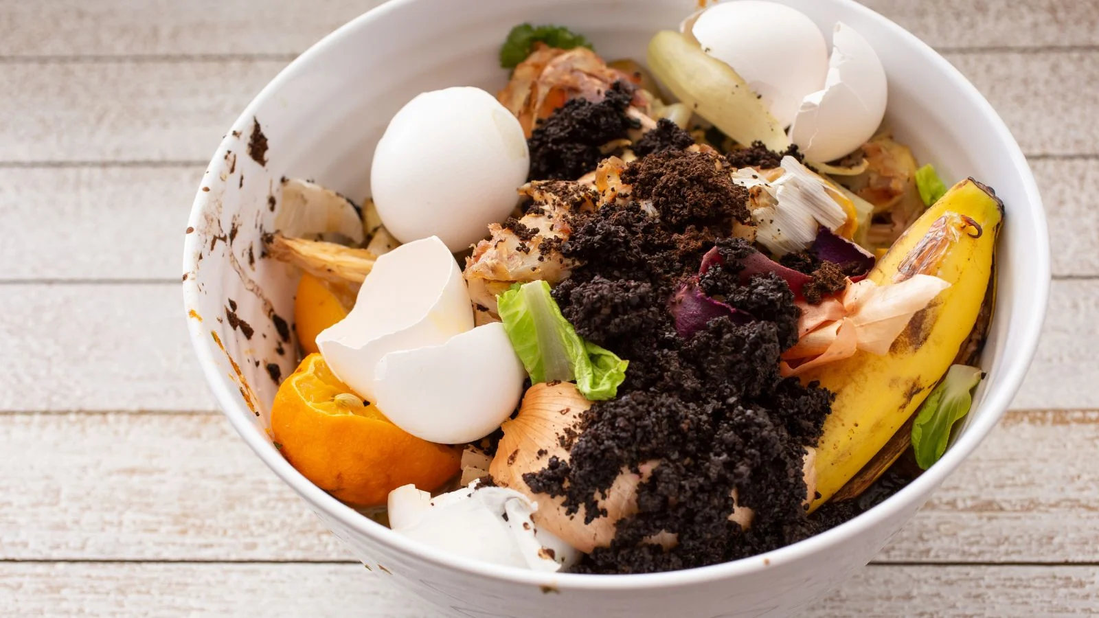 Миска з компостом, різноманітними кухонними відходами та іншими відкладеннями в білому контейнері на дерев'яній поверхні. Компостер Lomi переробляє більшість відходів.